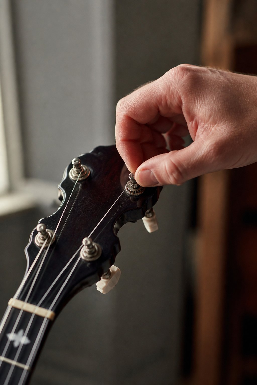 A hand turning a geared tuning peg on a banjo headstock, strings catching the light